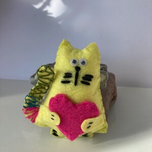 Puede incluir: Un gato de fieltro amarillo hecho a mano sosteniendo un corazón rosa. El gato tiene ojos saltones, bigotes negros y una nariz negra. Tiene una cola verde y multicolor. El gato es un artículo lindo, caprichoso y decorativo.
