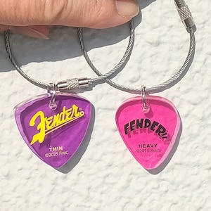 以下が含まれることがあります： 銀色のチェーンにFenderのギターピックが2つ付いています。左側のピックは紫色で、黄色のFenderロゴと「THIN ©2025 FMIC」の文字が書かれています。右側のピックはピンク色で、黒いFenderロゴと「HEAVY ©2025 FMIC」の文字が書かれています。