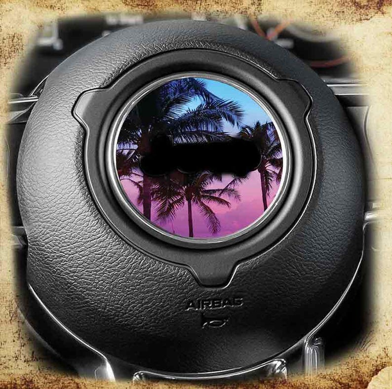 May include: Black steering wheel airbag cover with a circular insert featuring a tropical sunset scene with palm trees. The word "AIRBAG" is visible at the bottom of the cover.