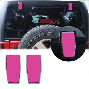May include: Two bright pink rectangular covers with rounded corners designed for vehicle rear window hinges. The image shows these covers installed on the black hinges of a vehicle's rear window, positioned above a spare tire and red brake light.