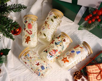 May include: A collection of glass jars with wooden lids, decorated with festive Christmas-themed illustrations. The jars feature designs like gingerbread men, snowmen, and gift boxes. The jars are arranged on a white surface with Christmas decorations.