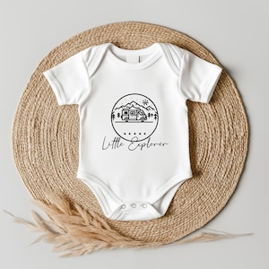 Könnte beinhalten: Ein weißer kurzärmeliger Babybody mit einem schwarzen Grafikdruck eines Wohnmobils, Bergen, Bäumen und einer Sonne in einem Kreis. Unter dem Kreis befinden sich fünf kleine Sterne und der Schriftzug Little Explorer in Schreibschrift.