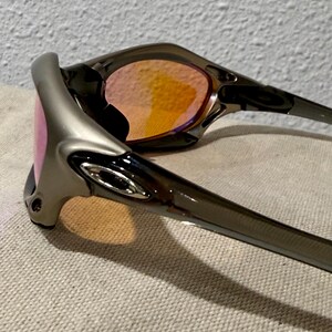 May include: A pair of sunglasses with a silver frame and tinted lenses. The lenses have a gradient of pink to orange. The sunglasses are resting on a textured, light-colored surface.