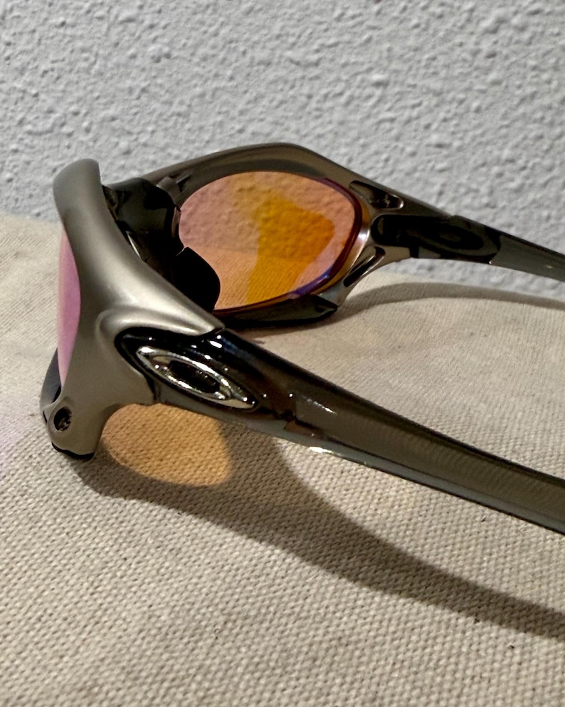 May include: A pair of sunglasses with a silver frame and tinted lenses. The lenses have a gradient of pink to orange. The sunglasses are resting on a textured, light-colored surface.