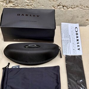 May include: A black Oakley sunglasses case, box, drawstring bag, cleaning cloth, and paperwork are displayed on a neutral surface. The box is labeled "OAKLEY" and "DESIGNED BY OAKLEY IN CALIFORNIA". The sunglasses case is zippered and has a metal Oakley logo.