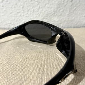 K&ouml;nnte beinhalten: Schwarze Sonnenbrille mit dunkelgrauen Gl&auml;sern. Die Sonnenbrille hat ein schlankes, modernes Design mit einem geschwungenen Rahmen und einem kleinen Metallakzent am B&uuml;gel. Die Gl&auml;ser sind get&ouml;nt und bieten Schatten und Schutz vor der Sonne.