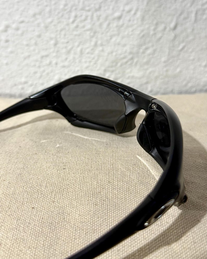 K&ouml;nnte beinhalten: Schwarze Sonnenbrille mit dunkelgrauen Gl&auml;sern. Die Sonnenbrille hat ein schlankes, modernes Design mit einem geschwungenen Rahmen und einem kleinen Metallakzent am B&uuml;gel. Die Gl&auml;ser sind get&ouml;nt und bieten Schatten und Schutz vor der Sonne.