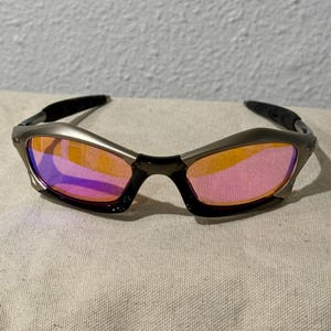 May include: Sport sunglasses with a silver frame and black temples. The lenses have a gradient of orange to pink hues. The sunglasses are resting on a neutral-colored fabric.