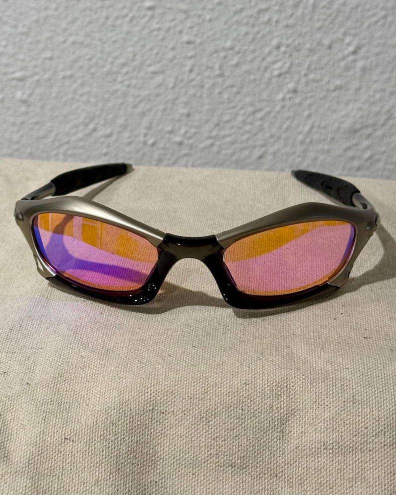 May include: Sport sunglasses with a silver frame and black temples. The lenses have a gradient of orange to pink hues. The sunglasses are resting on a neutral-colored fabric.