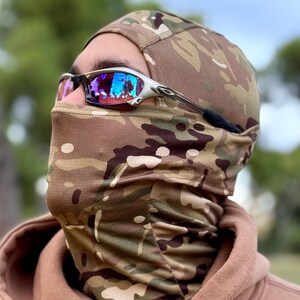 May include: A camouflage balaclava and face covering with a matching cap. The balaclava is made of a breathable fabric with a brown, green, and tan pattern. Silver-framed sunglasses with reflective lenses are worn over the balaclava.