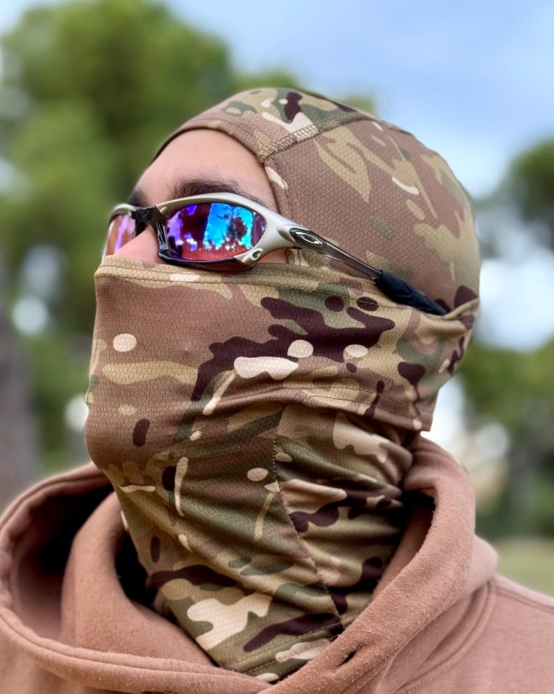 May include: A camouflage balaclava and face covering with a matching cap. The balaclava is made of a breathable fabric with a brown, green, and tan pattern. Silver-framed sunglasses with reflective lenses are worn over the balaclava.