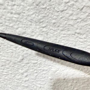K&ouml;nnte beinhalten: Nahaufnahme eines schwarzen Objekts mit dem Aufdruck "SPLICE". Das Objekt hat eine ovale Form und eine strukturierte Oberfl&auml;che. Der Hintergrund ist eine wei&szlig;e, strukturierte Wand.