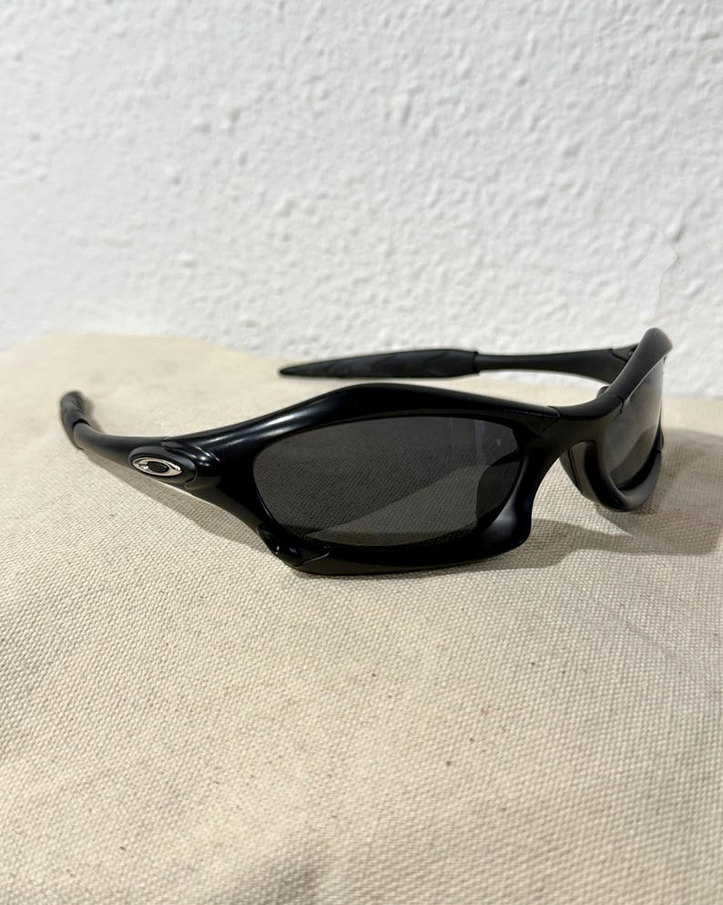 K&ouml;nnte beinhalten: Schwarze Sonnenbrille mit geschwungenem Rahmen und dunklen Gl&auml;sern. Die Sonnenbrille hat ein schlankes, modernes Design mit einem kleinen silbernen Logo an der Seite. Die B&uuml;gel umschlie&szlig;en die Ohren f&uuml;r einen sicheren Sitz.