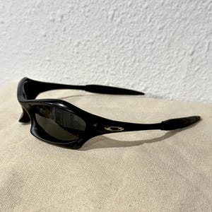 K&ouml;nnte beinhalten: Schwarze Sonnenbrille mit geschwungenem Design. Die Sonnenbrille hat dunkle Gl&auml;ser und eine gl&auml;nzende Oberfl&auml;che. Die B&uuml;gel sind d&uuml;nn und biegen sich um die Ohren. Das Oakley-Logo ist an der Seite des Rahmens sichtbar.