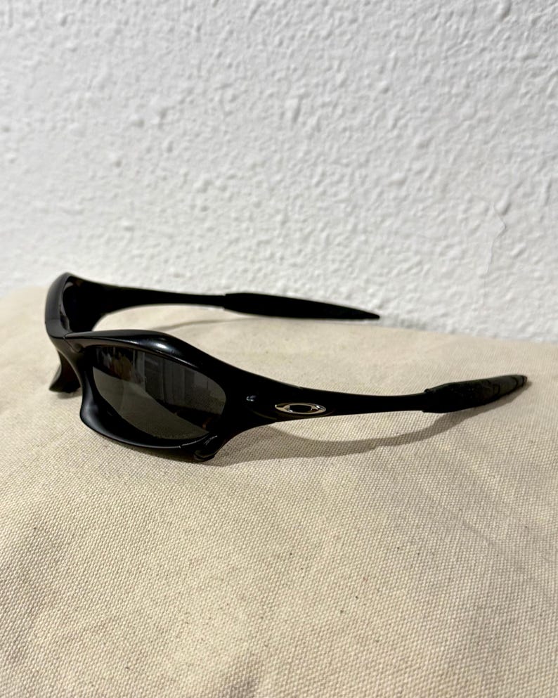 K&ouml;nnte beinhalten: Schwarze Sonnenbrille mit geschwungenem Design. Die Sonnenbrille hat dunkle Gl&auml;ser und eine gl&auml;nzende Oberfl&auml;che. Die B&uuml;gel sind d&uuml;nn und biegen sich um die Ohren. Das Oakley-Logo ist an der Seite des Rahmens sichtbar.