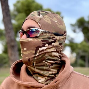 May include: A camouflage balaclava and neck gaiter set. The balaclava covers the head and neck, featuring a brown, green, and tan pattern. The person is wearing silver-framed sunglasses with red lenses. The background is a blurred outdoor scene.