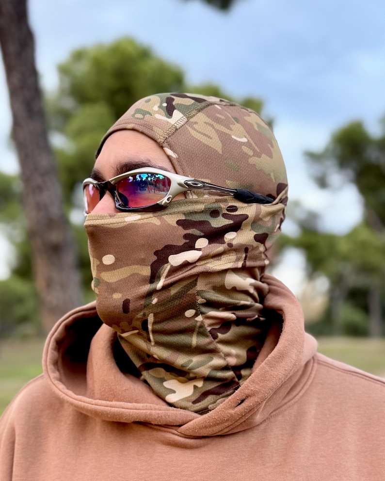 May include: A camouflage balaclava and neck gaiter set. The balaclava covers the head and neck, featuring a brown, green, and tan pattern. The person is wearing silver-framed sunglasses with red lenses. The background is a blurred outdoor scene.