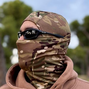 K&ouml;nnte beinhalten: Eine Tarn-Balaclava mit einem braunen, gr&uuml;nen und beigen Muster. Die Sturmhaube bedeckt Kopf und Hals, mit einer Gesichts&ouml;ffnung. Eine schwarze Sonnenbrille wird getragen. Die Person tr&auml;gt einen braunen Kapuzenpullover.