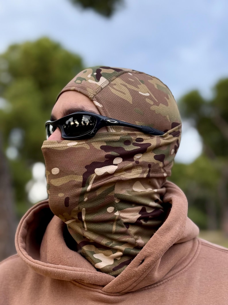 K&ouml;nnte beinhalten: Eine Tarn-Balaclava mit einem braunen, gr&uuml;nen und beigen Muster. Die Sturmhaube bedeckt Kopf und Hals, mit einer Gesichts&ouml;ffnung. Eine schwarze Sonnenbrille wird getragen. Die Person tr&auml;gt einen braunen Kapuzenpullover.