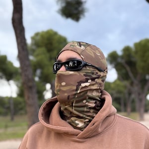 K&ouml;nnte beinhalten: Eine Tarn-Balaclava und ein Halstuch, kombiniert mit einer schwarzen Sonnenbrille und einem beigen Kapuzenpullover. Die Balaclava und das Halstuch weisen ein braunes, gr&uuml;nes und beiges Tarnmuster auf.