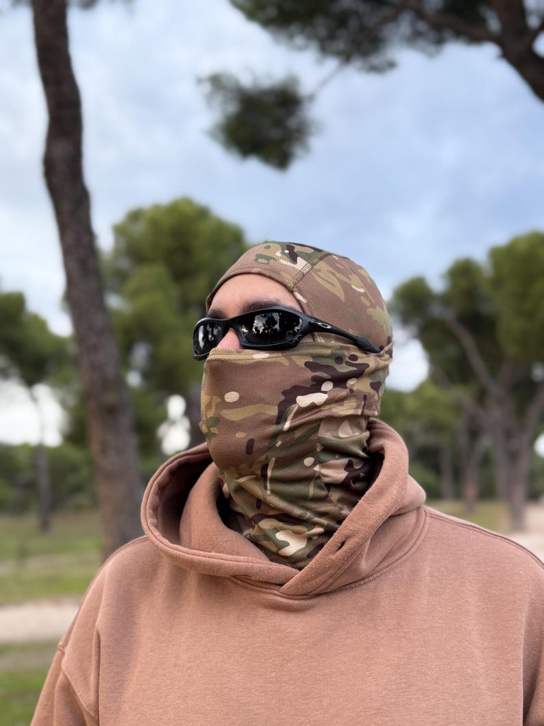 K&ouml;nnte beinhalten: Eine Tarn-Balaclava und ein Halstuch, kombiniert mit einer schwarzen Sonnenbrille und einem beigen Kapuzenpullover. Die Balaclava und das Halstuch weisen ein braunes, gr&uuml;nes und beiges Tarnmuster auf.