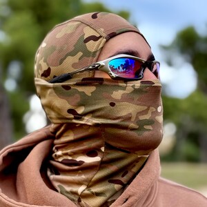 May include: A camouflage balaclava and neck gaiter set. The balaclava covers the head and face, with a neck gaiter extending down to the chest. Silver-framed sunglasses with blue lenses are worn over the balaclava.