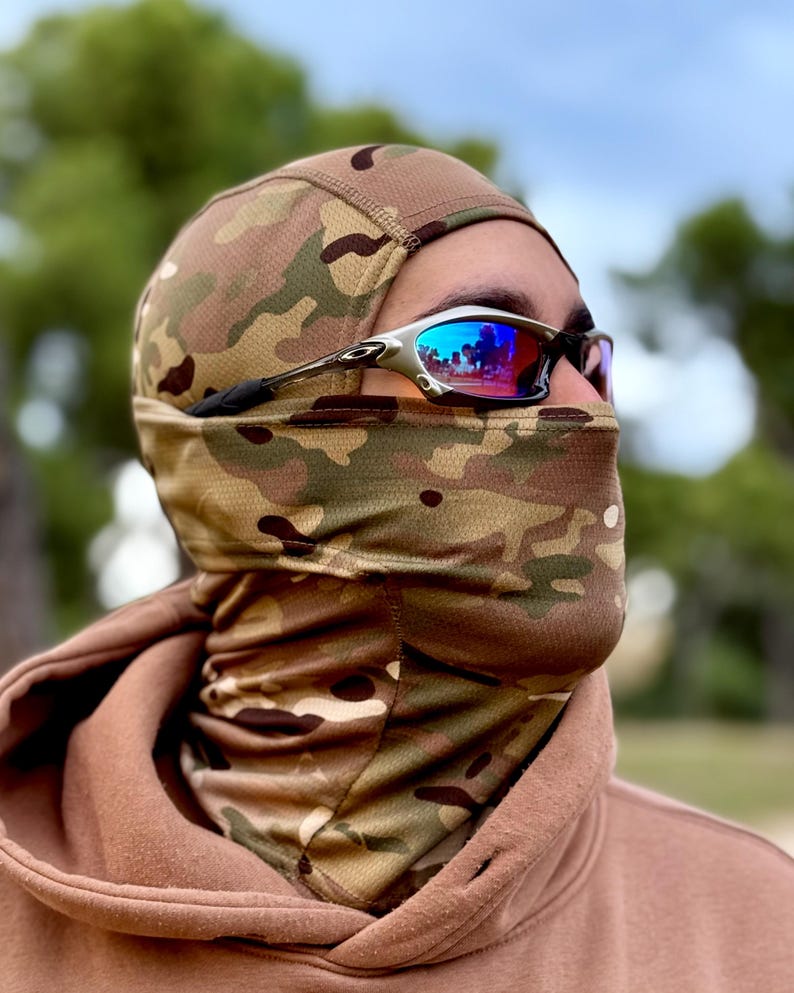 May include: A camouflage balaclava and neck gaiter set. The balaclava covers the head and face, with a neck gaiter extending down to the chest. Silver-framed sunglasses with blue lenses are worn over the balaclava.
