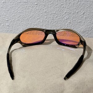 May include: Sport sunglasses with a dark gray frame and orange-tinted lenses. The arms are black and curve to fit behind the ears. The lenses have a slight purple hue at the bottom. The sunglasses are designed for outdoor activities.