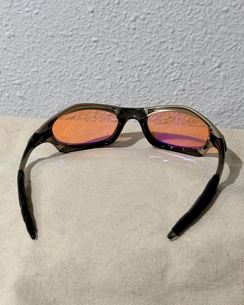 May include: Sport sunglasses with a dark gray frame and orange-tinted lenses. The arms are black and curve to fit behind the ears. The lenses have a slight purple hue at the bottom. The sunglasses are designed for outdoor activities.