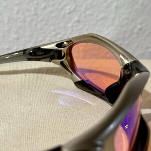May include: Close-up of a pair of sunglasses with a silver frame and orange-tinted lenses. The lenses have a slight purple hue. The sunglasses are angled, showing the details of the frame and lens shape.