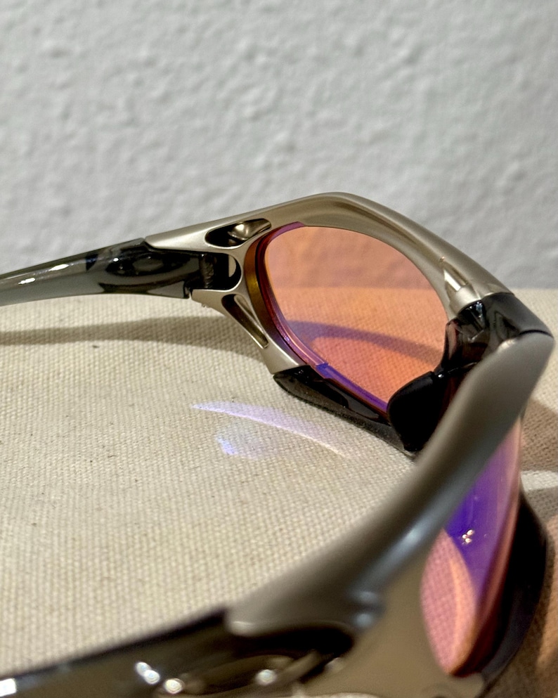 May include: Close-up of a pair of sunglasses with a silver frame and orange-tinted lenses. The lenses have a slight purple hue. The sunglasses are angled, showing the details of the frame and lens shape.