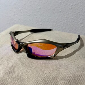 May include: Sport sunglasses with a silver frame and black temples. The lenses are a gradient of orange and pink. The sunglasses are resting on a beige surface.