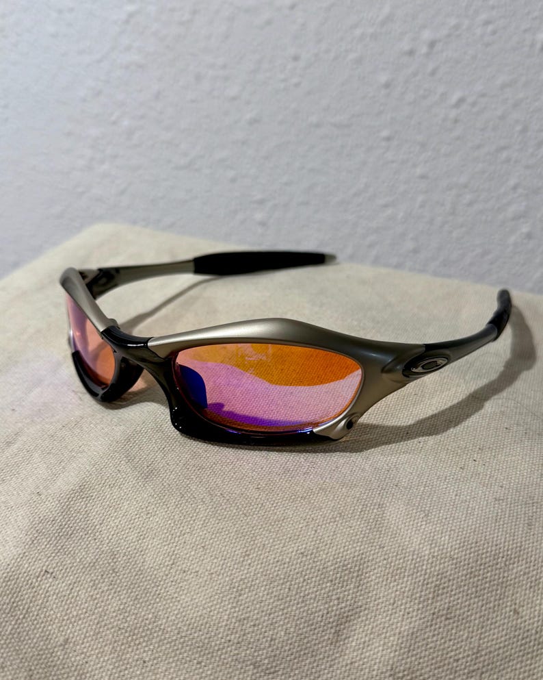 May include: Sport sunglasses with a silver frame and black temples. The lenses are a gradient of orange and pink. The sunglasses are resting on a beige surface.