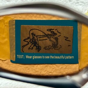 May include: A close-up of a pair of glasses with a blue and brown rectangular sticker. The sticker features a drawing of a person fishing and the text: "TEST; Wear glasses to see the beautiful pattern."