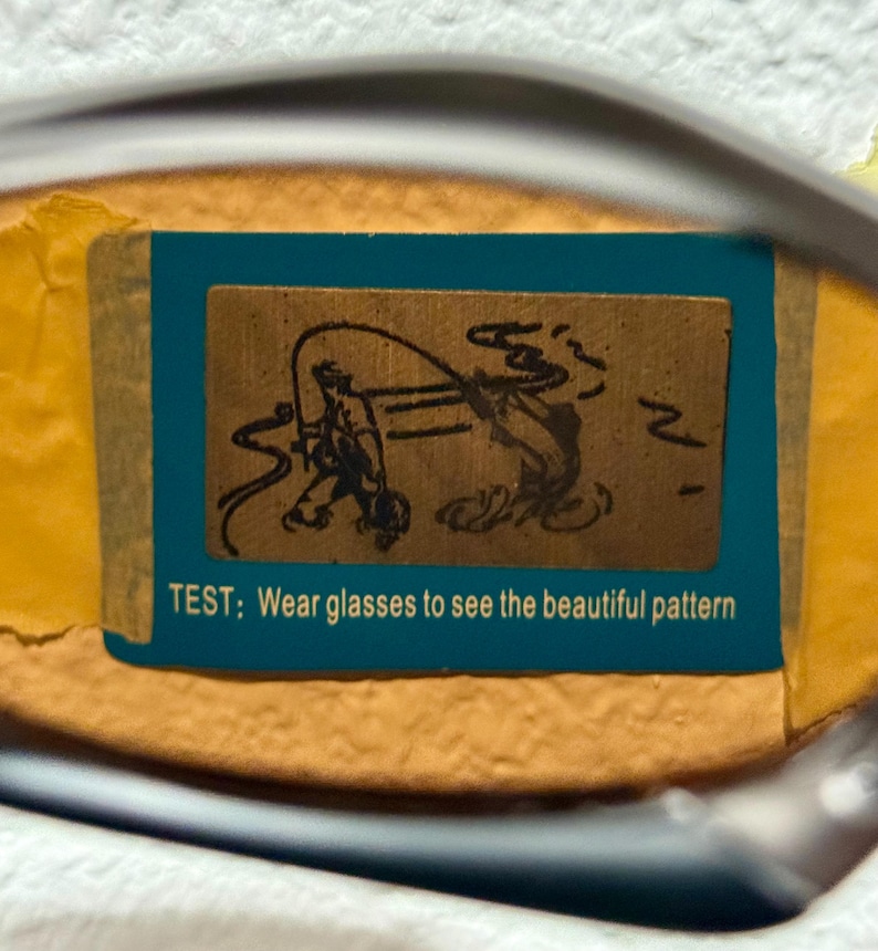 May include: A close-up of a pair of glasses with a blue and brown rectangular sticker. The sticker features a drawing of a person fishing and the text: "TEST; Wear glasses to see the beautiful pattern."