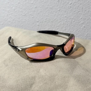 May include: Sport sunglasses with a metallic gray frame and black temples. The lenses are a gradient of pink and orange. The sunglasses are resting on a neutral-colored fabric.