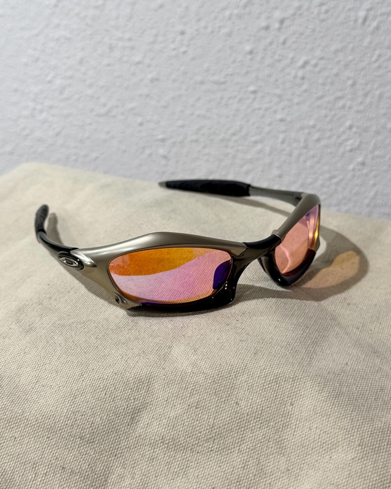 May include: Sport sunglasses with a metallic gray frame and black temples. The lenses are a gradient of pink and orange. The sunglasses are resting on a neutral-colored fabric.