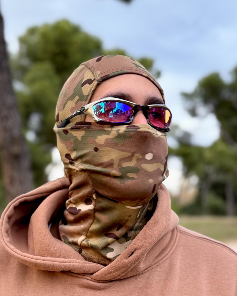 May include: A person wearing a camouflage balaclava and a brown hoodie. The person is also wearing sunglasses with a silver frame and reflective lenses. The balaclava covers the head and neck, with a camouflage pattern in shades of brown, green, and tan.