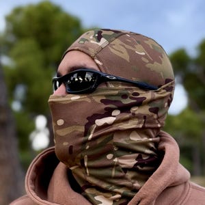 K&ouml;nnte beinhalten: Eine Tarn-Balaclava und eine schwarze Sonnenbrille. Die Sturmhaube hat ein Muster aus Braun, Gr&uuml;n und Beige. Die Sonnenbrille hat einen schwarzen Rahmen. Die Person tr&auml;gt einen braunen Kapuzenpullover.