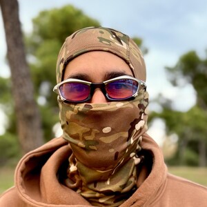 May include: A person wearing a camouflage balaclava, matching neck gaiter, and brown hoodie. Silver-framed sunglasses with red lenses are worn over the balaclava. The background features trees and a cloudy sky.