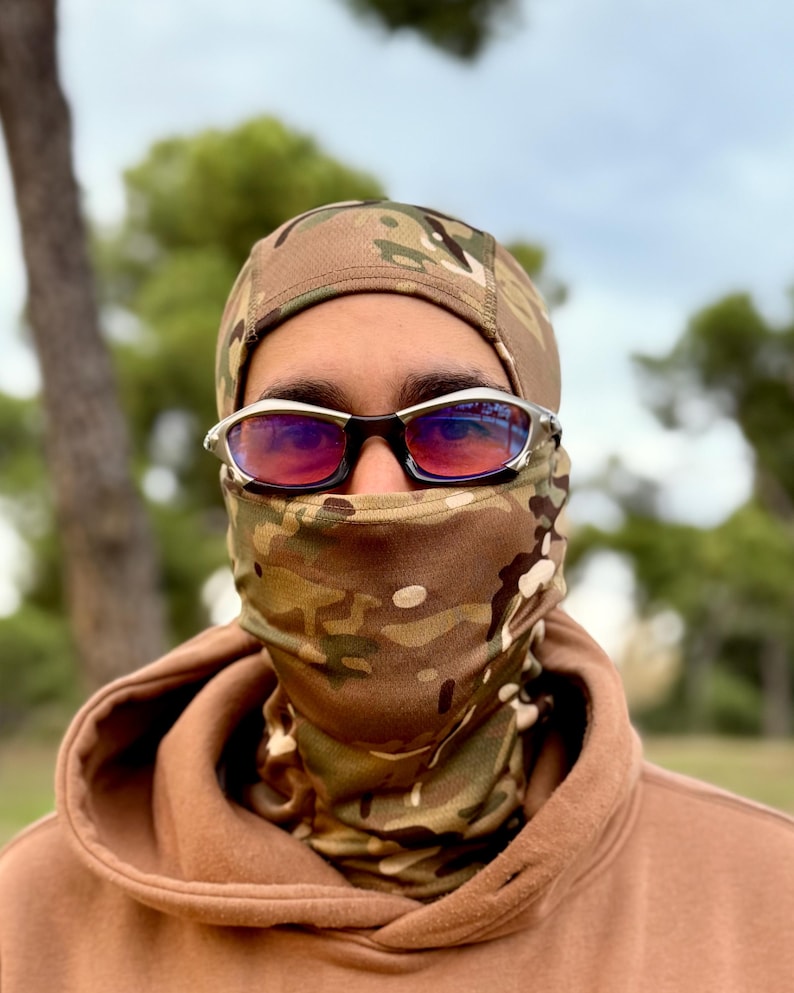 May include: A person wearing a camouflage balaclava, matching neck gaiter, and brown hoodie. Silver-framed sunglasses with red lenses are worn over the balaclava. The background features trees and a cloudy sky.