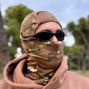 K&ouml;nnte beinhalten: Eine Tarn-Balaclava und ein Halstuch mit passendem Muster aus Gr&uuml;n, Braun und Beige. Die Person tr&auml;gt eine schwarze Sonnenbrille und einen braunen Kapuzenpullover. Die Sturmhaube bedeckt Kopf und Hals, das Gesicht ist frei.