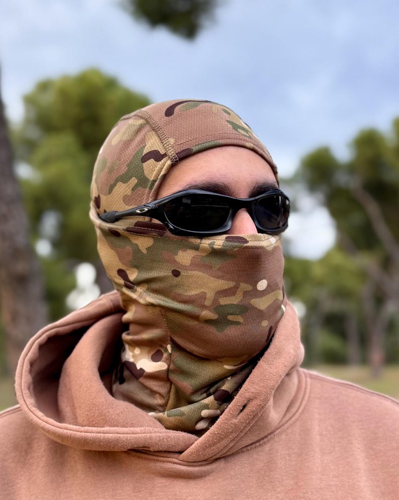 K&ouml;nnte beinhalten: Eine Tarn-Balaclava und ein Halstuch mit passendem Muster aus Gr&uuml;n, Braun und Beige. Die Person tr&auml;gt eine schwarze Sonnenbrille und einen braunen Kapuzenpullover. Die Sturmhaube bedeckt Kopf und Hals, das Gesicht ist frei.