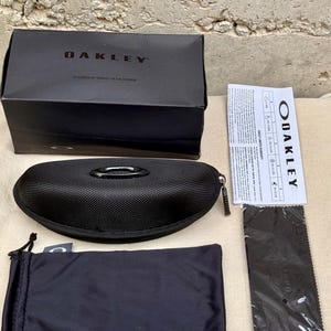 K&ouml;nnte beinhalten: Gezeigt werden ein schwarzes Oakley-Sonnenbrillenetui, eine Box und Zubeh&ouml;r. Auf der Box steht "OAKLEY" und "DESIGNED BY OAKLEY IN CALIFORNIA". Enthalten sind ein Etui mit Rei&szlig;verschluss, ein Kordelzugbeutel, ein Reinigungstuch und eine Garantiekarte.