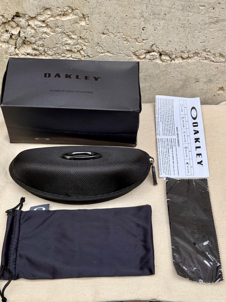K&ouml;nnte beinhalten: Gezeigt werden ein schwarzes Oakley-Sonnenbrillenetui, eine Box und Zubeh&ouml;r. Auf der Box steht "OAKLEY" und "DESIGNED BY OAKLEY IN CALIFORNIA". Enthalten sind ein Etui mit Rei&szlig;verschluss, ein Kordelzugbeutel, ein Reinigungstuch und eine Garantiekarte.
