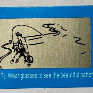 K&ouml;nnte beinhalten: Eine blaue rechteckige Karte mit einem silbernen Rechteck in der Mitte. Das silberne Rechteck hat eine schwarze Illustration einer angelnden Person. Der Text unten lautet: "TEST: Tragen Sie eine Brille, um das sch&ouml;ne Muster zu sehen."