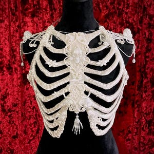 Peut inclure: Un bustier blanc en forme de cage thoracique, orné de motifs floraux et d'un crâne. Le bustier est présenté sur un mannequin noir, sur fond de velours rouge. Un petit pendentif en forme de main est suspendu.