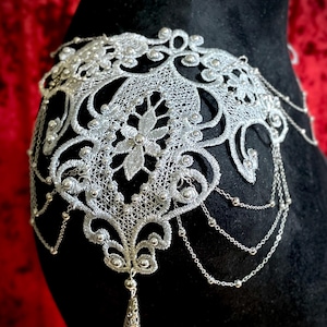 Peut inclure: Coiffe argentée en dentelle avec des motifs floraux et géométriques. Le design complexe présente des chaînes pendantes et des perles. La coiffe est présentée sur une surface noire avec un fond rouge, mettant en valeur ses détails délicats.