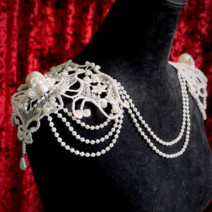 May include: Ivory lace shoulder piece with pearl bead strands and a skull accent. The shoulder piece is displayed on a black mannequin, against a red velvet backdrop. The lace design features floral and scroll patterns.