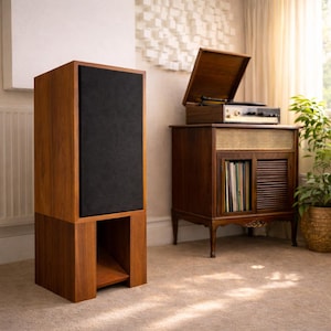 Può includere: Un grande altoparlante in legno con un pannello frontale in tessuto nero. L'altoparlante fa parte di un'installazione audio vintage, che include un giradischi e un mobile con spazio per i dischi. L'installazione si trova in una stanza con una combinazione di colori neutri.